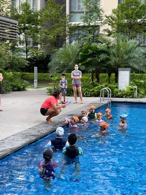 Coach Jina crouching poolside with a clipboard, briefing a group of kids in the water